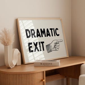 May include: A framed print featuring the text "DRAMATIC EXIT" in black, with a hand pointing right. The artwork is on a wooden table, with a vase and a book titled "MATISSE". The overall aesthetic is minimalist.