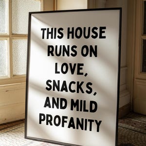 May include: A rectangular sign with a black frame and a white background. The sign features the text "THIS HOUSE RUNS ON LOVE, SNACKS, AND MILD PROFANITY" in bold, black letters. The sign is displayed indoors, near a window.