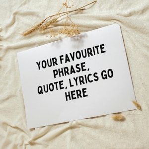 Op de afbeelding: Een witte rechthoekige kaart met de zwarte tekst "YOUR FAVOURITE PHRASE, QUOTE, LYRICS GO HERE". De kaart ligt op een beige, getextureerd oppervlak, met gedroogde planten op de achtergrond.