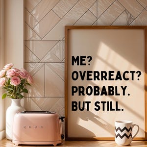 Pode incluir: Uma impressão emoldurada com o texto "ME? OVERREACT? PROBABLY. BUT STILL." em letras pretas sobre um fundo bege. Uma torradeira rosa e uma caneca com padrão preto e branco estão em um balcão na frente da impressão.