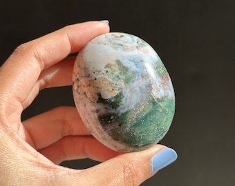 May include: A smooth, polished green, white and brown agate stone with a swirling pattern. The stone is oval shaped and is being held in a hand.