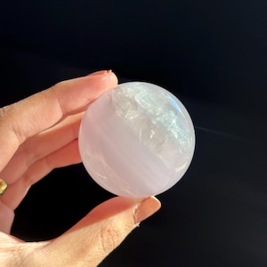 May include: A translucent, light pink sphere with internal crystalline structures. The sphere is held against a dark background, showcasing its polished surface and subtle color variations. The object appears to be a decorative crystal or gemstone.