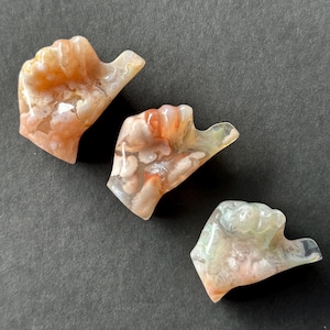 May include: Three hand-shaped agate stones in shades of orange, white, and clear. The stones have a polished, translucent appearance, with visible patterns and inclusions. The thumbs-up gesture is clearly defined in each stone.