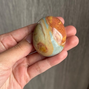 May include: A smooth, polished, egg-shaped stone featuring a marbled pattern with distinct bands of burnt orange, cream, tan, and soft sage green. The surface has a glossy finish that reflects light, highlighting the natural color variations.
