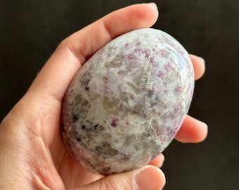 May include: A smooth, white and grey egg-shaped crystal with pink and red inclusions. The crystal is being held in a hand.