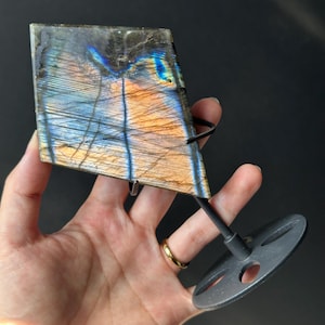 Könnte beinhalten: Ein polierter Labradorit-Edelstein in Rhombusform mit schillernden blauen, orangefarbenen und goldenen Farbtönen. Er wird von einem schwarzen Metallständer gehalten. Der Hintergrund ist dunkel.