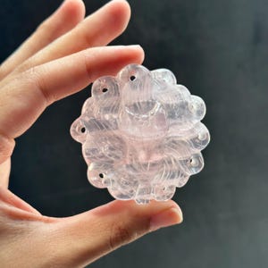 May include: A carved rose quartz pendant, held in a hand, features a detailed floral design. The translucent pink stone has multiple holes for stringing. The pendant's intricate carving and soft color create an elegant, natural aesthetic.