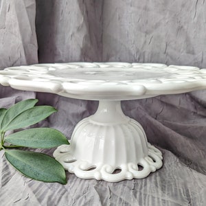 May include: A white, vintage-style cake stand with a scalloped edge and a pedestal base. The stand is made of ceramic or porcelain and features a decorative, openwork design around the base. The stand is approximately 10 inches in diameter.