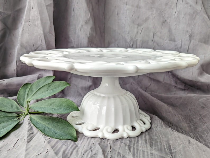 May include: A white, vintage-style cake stand with a scalloped edge and a pedestal base. The stand is made of ceramic or porcelain and features a decorative, openwork design around the base. The stand is approximately 10 inches in diameter.