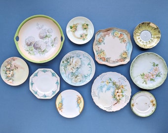 May include: A collection of 11 vintage porcelain plates with floral designs. The plates are in various sizes and shapes, and feature different floral patterns, including roses, hydrangeas, and other delicate blooms. The plates are all white with gold trim and are in excellent condition.