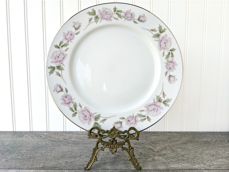 May include: A white porcelain dinner plate with a gold rim and a delicate floral design featuring pink roses and green leaves.