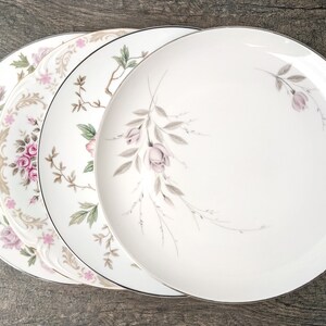 May include: A set of four white ceramic dinner plates with a delicate floral design featuring pink roses and grey leaves. The plates have a silver rim.