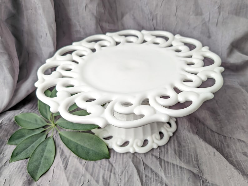 May include: A white, ornate cake stand with a scalloped edge and a pedestal base. The stand has a detailed, openwork design. A green leaf is placed on the side of the stand against a gray background.