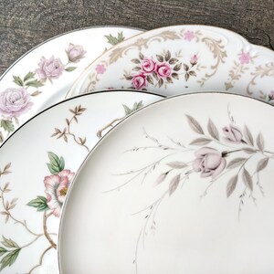 May include: Three white ceramic plates with floral designs. The top plate has a gold trim and a pink and brown floral pattern. The middle plate has a pink and green floral pattern. The bottom plate has a light pink and grey floral pattern.