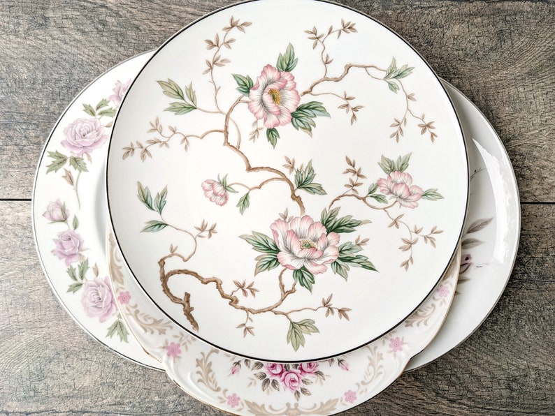 May include: Three white ceramic plates with a floral design. The top plate has a larger floral design with pink flowers and green leaves. The bottom two plates have a smaller floral design with pink flowers and green leaves.