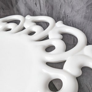 May include: A white ceramic serving tray with a decorative, openwork border. The tray has a glossy finish and a rectangular shape. The border features a series of curved, loop-like designs. The background is a textured gray fabric.