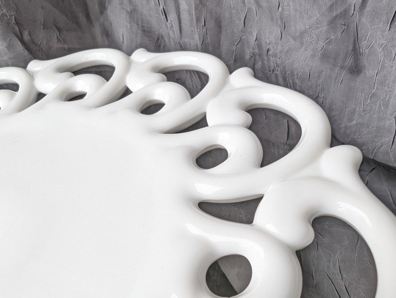 May include: A white ceramic serving tray with a decorative, openwork border. The tray has a glossy finish and a rectangular shape. The border features a series of curved, loop-like designs. The background is a textured gray fabric.