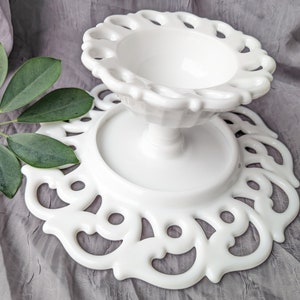 May include: A white, vintage-style pedestal cake stand with a decorative, openwork design. The stand features a round base and a scalloped edge, perfect for displaying desserts or other items. The stand is approximately 8 inches in diameter and 6 inches tall.