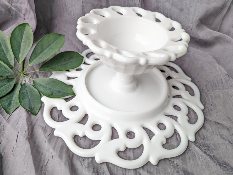 May include: A white, vintage-style pedestal cake stand with a decorative, openwork design. The stand features a round base and a scalloped edge, perfect for displaying desserts or other items. The stand is approximately 8 inches in diameter and 6 inches tall.