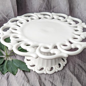 May include: A white, ornate cake stand with a decorative, openwork design around the edge. The stand has a circular top and a pedestal base. The stand is made of ceramic or porcelain and is suitable for displaying cakes or other desserts.