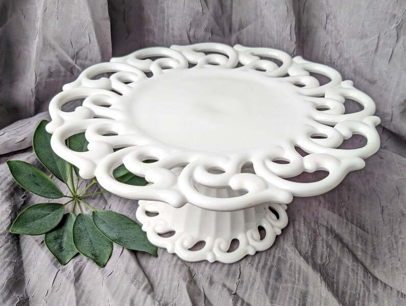 May include: A white, ornate cake stand with a decorative, openwork design around the edge. The stand has a circular top and a pedestal base. The stand is made of ceramic or porcelain and is suitable for displaying cakes or other desserts.