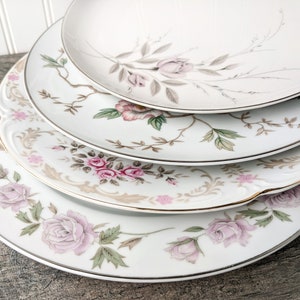 May include: Four white ceramic dinner plates with a floral design featuring pink roses and green leaves. The plates have a gold trim around the edges.