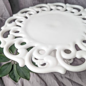 May include: A white, decorative plate with an ornate, openwork design around the edge. The plate has a smooth, glossy finish and a slightly raised center. The plate is set against a gray fabric background with green leaves.