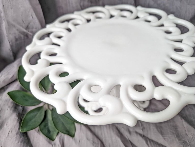 May include: A white, decorative plate with an ornate, openwork design around the edge. The plate has a smooth, glossy finish and a slightly raised center. The plate is set against a gray fabric background with green leaves.