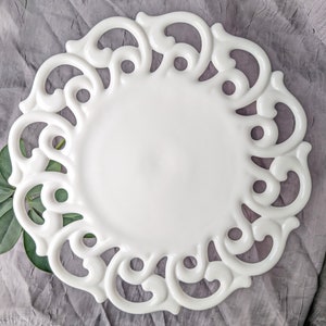 May include: A white ceramic plate with an ornate, openwork border. The plate has a smooth, glossy surface and a decorative, scroll-like design around the edge. Green leaves are placed near the plate. The plate is on a gray surface.