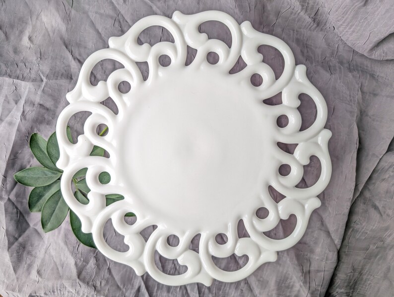 May include: A white ceramic plate with an ornate, openwork border. The plate has a smooth, glossy surface and a decorative, scroll-like design around the edge. Green leaves are placed near the plate. The plate is on a gray surface.
