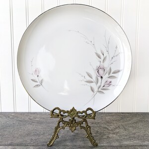 May include: A white ceramic dinner plate with a delicate floral design featuring pink roses and grey leaves. The plate has a gold rim and is displayed on a gold metal stand.