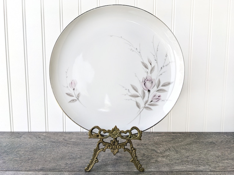 May include: A white ceramic dinner plate with a delicate floral design featuring pink roses and grey leaves. The plate has a gold rim and is displayed on a gold metal stand.