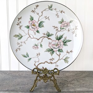 May include: A white porcelain plate with a gold rim features a floral design of pink and green flowers on a white background. The plate is displayed on a gold metal stand.