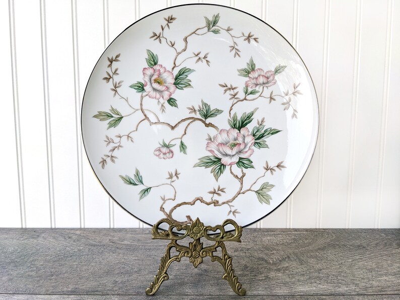 May include: A white porcelain plate with a gold rim features a floral design of pink and green flowers on a white background. The plate is displayed on a gold metal stand.