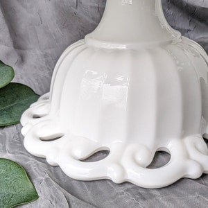 May include: A close-up of a white, ceramic pedestal cake stand. The stand features a scalloped edge with decorative cut-out designs. The base has a fluted design. The stand is on a gray surface with green leaves.