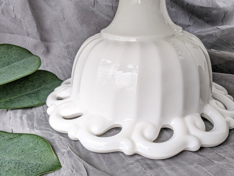 May include: A close-up of a white, ceramic pedestal cake stand. The stand features a scalloped edge with decorative cut-out designs. The base has a fluted design. The stand is on a gray surface with green leaves.
