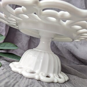 May include: A white, ornate cake stand with a scalloped edge and a pedestal base. The stand features a delicate, openwork design on the plate and base, perfect for displaying baked goods. The stand is made of ceramic or porcelain.