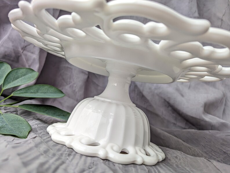 May include: A white, ornate cake stand with a scalloped edge and a pedestal base. The stand features a delicate, openwork design on the plate and base, perfect for displaying baked goods. The stand is made of ceramic or porcelain.