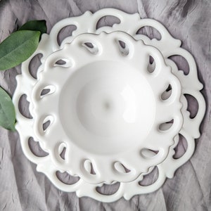 May include: Two white ceramic plates, one nested inside the other, with a delicate, openwork design around the edges. The plates are on a gray, textured surface, with green leaves in the upper left corner.