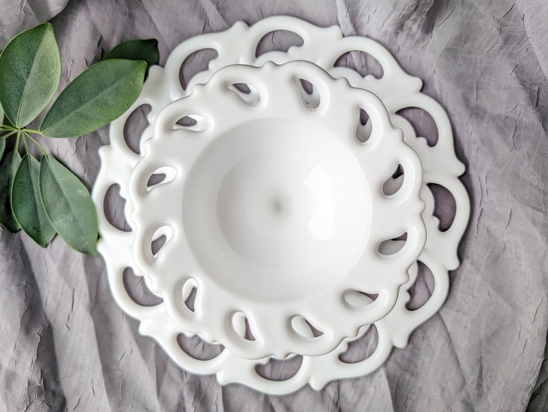 May include: Two white ceramic plates, one nested inside the other, with a delicate, openwork design around the edges. The plates are on a gray, textured surface, with green leaves in the upper left corner.