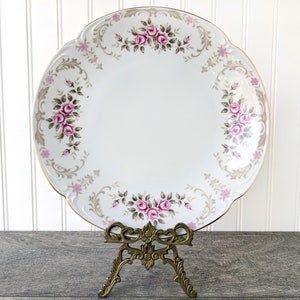 May include: A white porcelain serving platter with a gold rim and a floral design featuring pink and grey roses. The platter is on a gold metal stand.