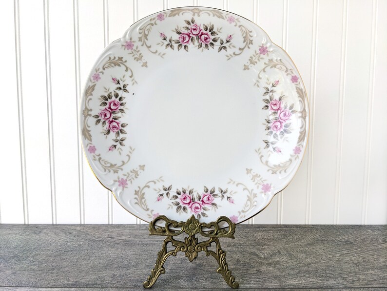 May include: A white porcelain serving platter with a gold rim and a floral design featuring pink and grey roses. The platter is on a gold metal stand.
