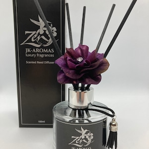 May include: A 100 ml glass bottle reed diffuser with a black label that says "JK-AROMAS luxury fragrances" and "Plum, Rose & Patchouli". The bottle has a silver cap and a purple flower with a rhinestone on top.  Black reeds are in the bottle.