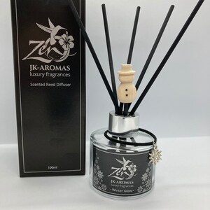 May include: A black box with white text that reads "JK-AROMAS luxury fragrances Scented Reed Diffuser 100ml". A clear glass bottle with a black lid and a silver band around the neck. The bottle is filled with a clear liquid and has a black ribbon tied around the neck. The bottle has a silver snowflake charm hanging from it. The bottle is sitting on a white surface. There are black reeds in the bottle. A wooden snowman ornament is on top of the reeds.