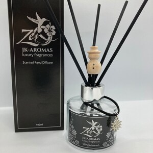 May include: A black and silver glass bottle with a black label that reads "JK-AROMAS luxury fragrances Gingerbread." The bottle has a silver cap and a wooden snowman-shaped stopper.  Black reed diffusers are in the bottle.  The bottle is sitting on a white surface.