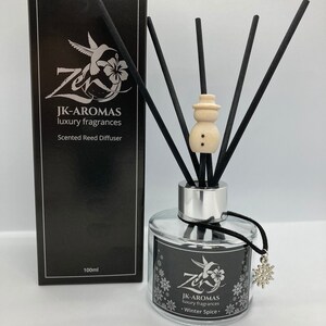 May include: A black and silver reed diffuser bottle with a snowman-shaped diffuser stick. The bottle has a black label with the text "JK-AROMAS luxury fragrances Scented Reed Diffuser 100ml" and a snowflake charm. The bottle is sitting on a white surface.