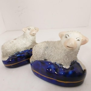 May include: Two ceramic sheep figurines. The sheep have white bodies with a textured, glittery appearance. They rest on a base of glossy, deep blue with a gold trim. The sheep have pink ears and a smiling expression.