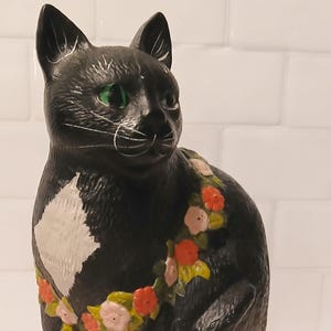 May include: A black cat figurine with a white chest patch and white paws. The cat has green eyes and is wearing a garland of small pink and orange flowers with green leaves around its neck. The surface of the figurine has a textured finish.
