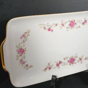 May include: A rectangular white porcelain serving tray featuring a delicate floral pattern of pink roses and green leaves arranged in garlands along the edges. The tray has integrated handles on both ends and a thin gold trim along the rim.