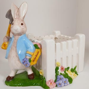 May include: A ceramic Peter Rabbit figurine holding a shovel and a basket with the text "Peter Rabbit". The figurine is wearing a blue jacket and stands next to a white picket fence planter with colorful flowers.